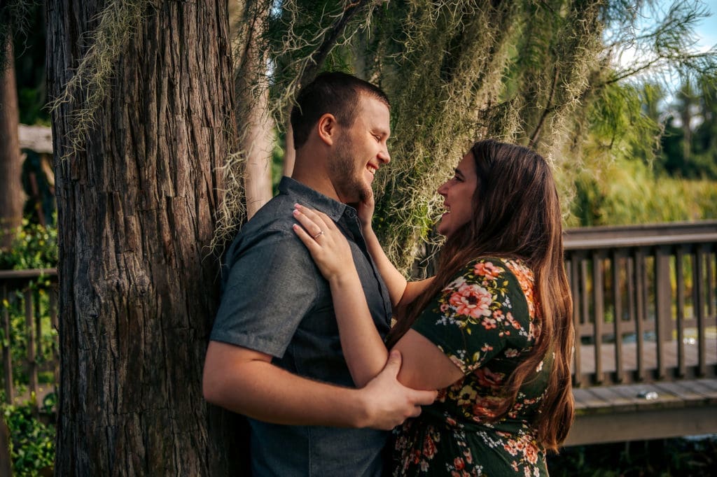 Engaged couple leaning against tree at Knotted Roots on the Lake